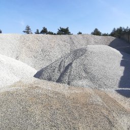 Star Budowa Inwestycje Sp z o.o. - Góra jasnego kruszywa pod błękitnym niebem, widok z dołu. Na szczycie w oddali widoczne drzewa. Kruszywo o różnej frakcji i odcieniach szarości.