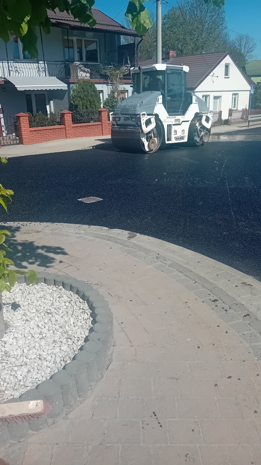 Walec drogowy HAMM ubija świeżo wylany asfalt na ulicy, widoczne domy w tle. Obrzeże chodnika z kostki brukowej i białego kamienia.