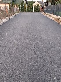 Wykończona, asfaltowa droga dojazdowa z drobnym żwirem po bokach, prowadząca w kierunku domu z zielonymi tujami. Widok z perspektywy wjazdu.