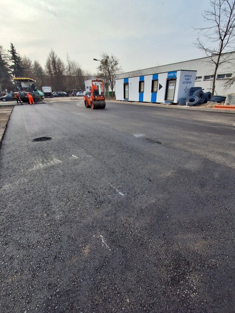 Walec drogowy ubija świeżo wylaną, czarną nawierzchnię asfaltową na drodze. W tle budynek modułowy i pracownicy przy maszynie do asfaltu.