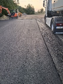 Fragment nowo położonej, czarnej nawierzchni asfaltowej, z widocznym obrzeżem, ciężarówką i sprzętem budowlanym w tle, w słoneczny dzień.
