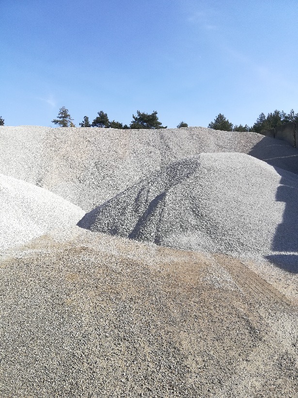 Góra jasnego kruszywa pod błękitnym niebem, widok z dołu. Na szczycie w oddali widoczne drzewa. Kruszywo o różnej frakcji i odcieniach szarości.