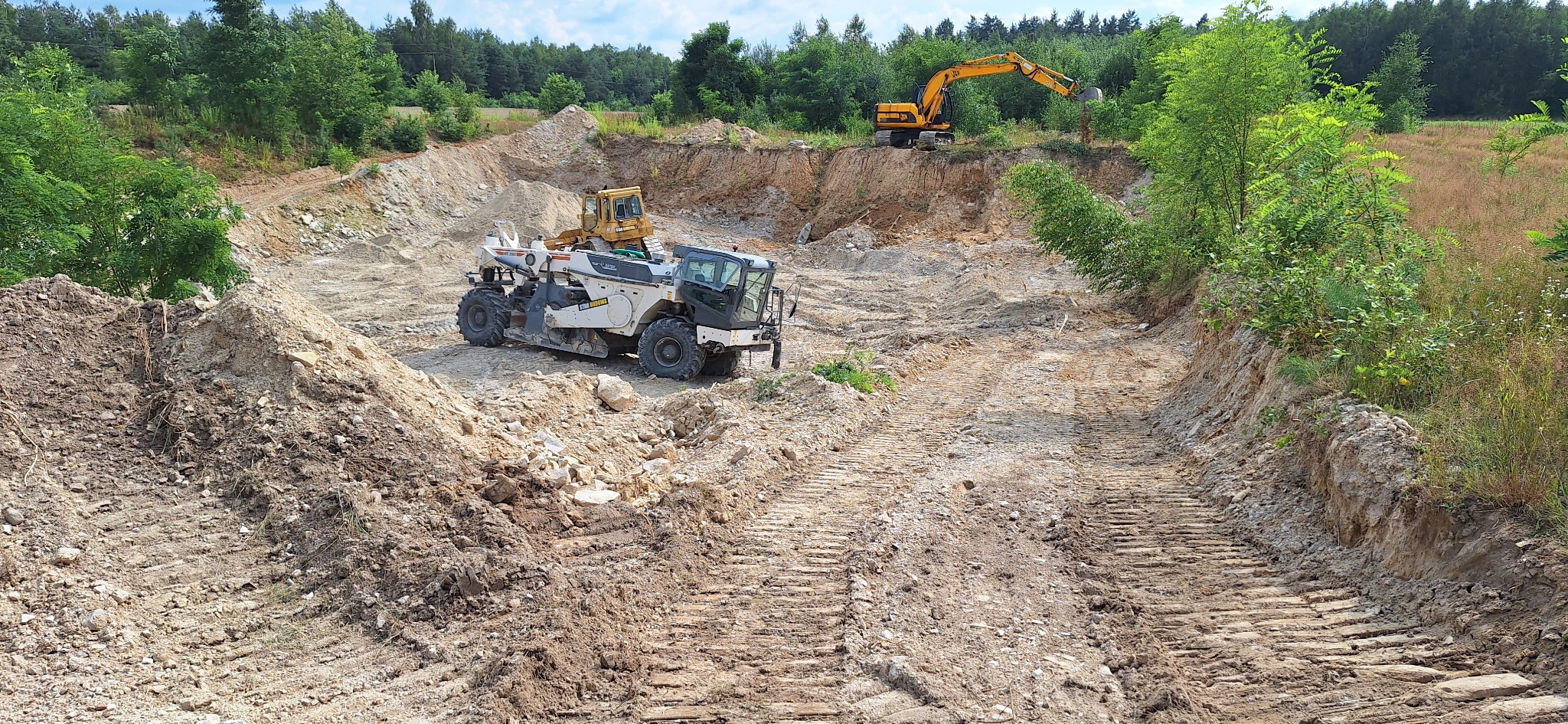 Krajobraz po niwelacji terenu z widocznymi śladami maszyn, koparką w tle i wzgórzem ziemi na pierwszym planie. Prace ziemne w otoczeniu zieleni.