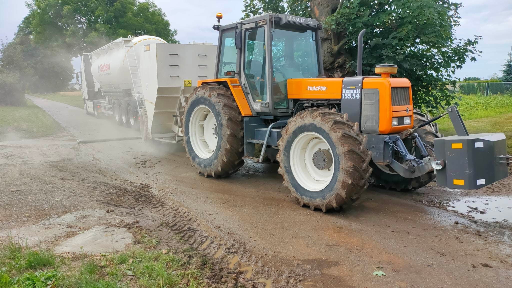 Pomarańczowy traktor Renault 155.54 z cysterną w trakcie prac polowych, rozpylający substancję na ziemi. Widoczne ślady opon na błotnistej drodze.
