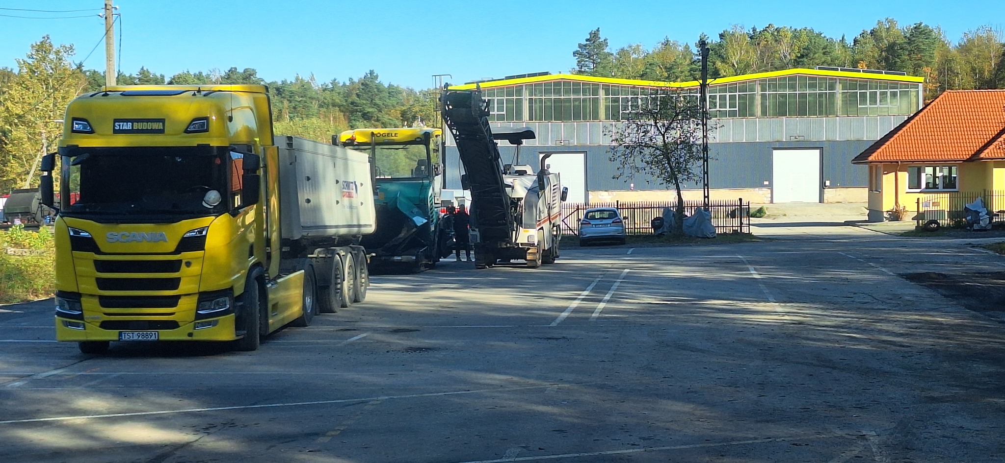 Żółty ciągnik Scania z naczepą i maszyny do budowy dróg (Vögele) na placu budowy. W tle budynek z żółtymi akcentami i drzewa. Słoneczny dzień.