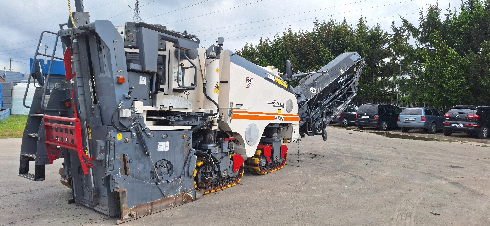 Frezerka Wirtgen W 100 F na gąsienicach, zaparkowana na placu, z zielenią w tle. Maszyna do frezowania nawierzchni asfaltowych.