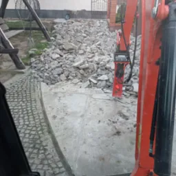 Widok z kabiny na gruzowisko po wyburzeniu, z fragmentem pomarańczowego ramienia koparki z młotem pneumatycznym. W tle brama z ceglanymi słupkami.