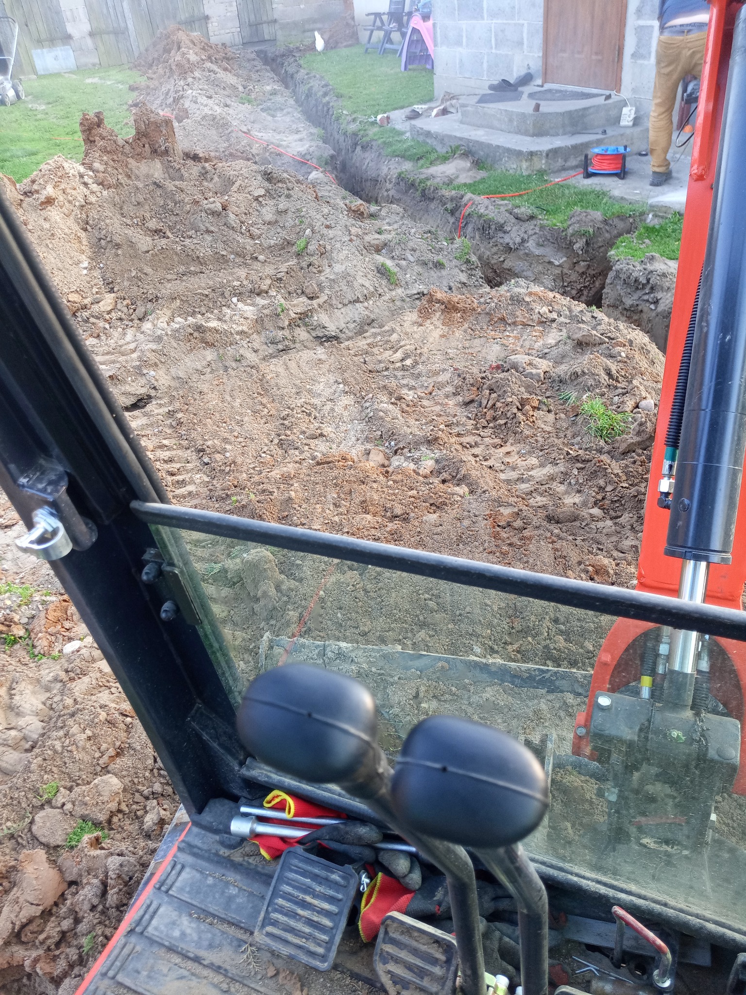 Wykopany rów w ziemi, widok z kabiny małej koparki. Na ziemi ślady opon, w tle budynek z betonowych pustaków i elementy instalacji.