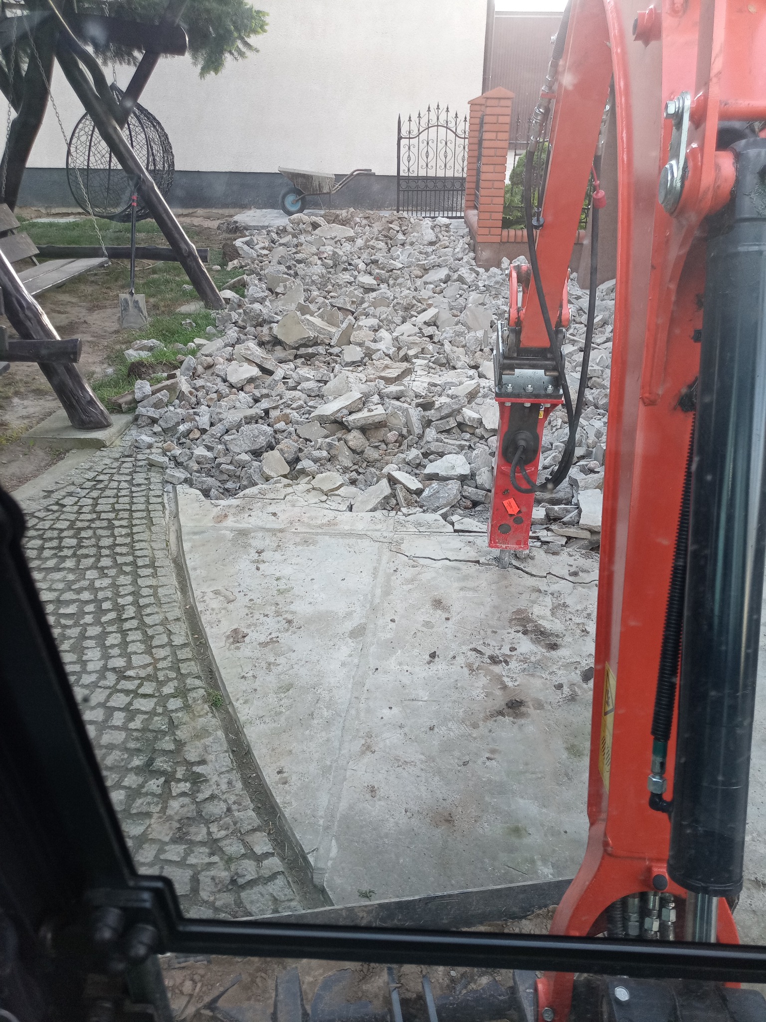 Widok z kabiny na gruzowisko po wyburzeniu, z fragmentem pomarańczowego ramienia koparki z młotem pneumatycznym. W tle brama z ceglanymi słupkami.