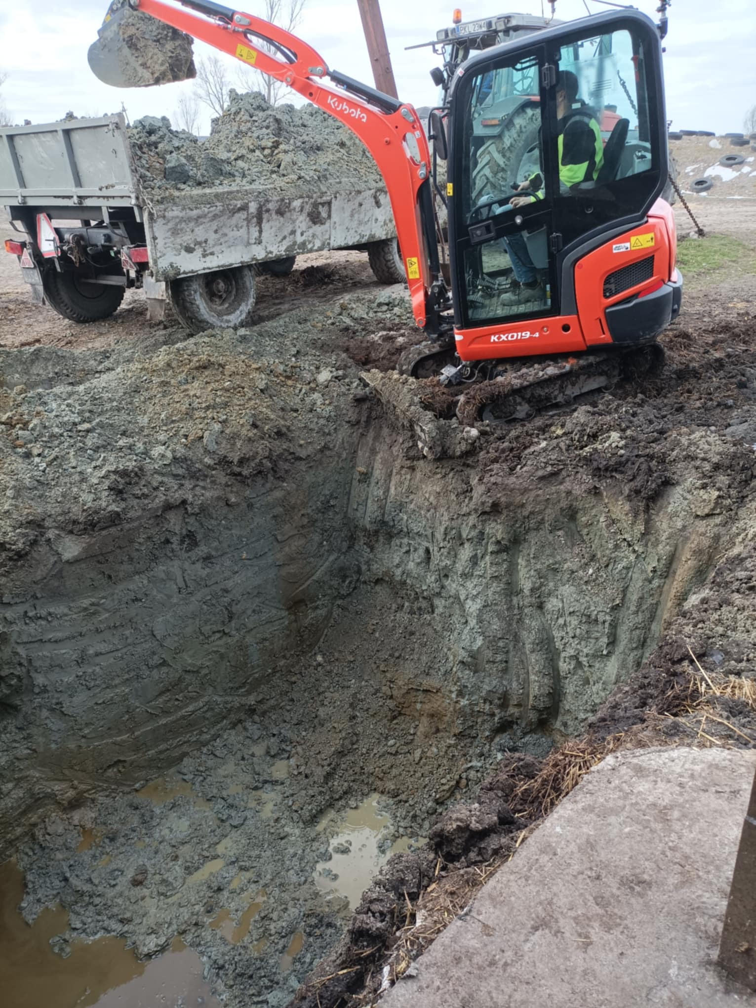 Pomarańczowa koparka Kubota KX019-4 nabiera ziemię do przyczepy podczas wykopu, widoczny operator w kabinie i głęboki wykop z wodą na dnie.