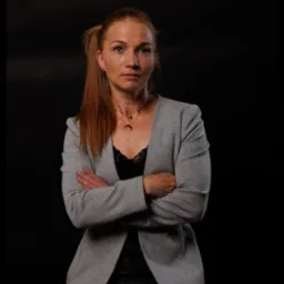 Portret kobiety w szarym żakiecie na czarnym tle, z rękami skrzyżowanymi, sugerujący profesjonalizm i pewność siebie, idealny na profil trenera personalnego.