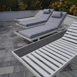 Rafał Macina - Nowoczesne, białe leżaki z szarymi poduszkami na szarym bruku, otoczone zielenią. Minimalistyczny design i spokojna kolorystyka tworzą relaksującą atmosferę.