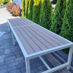 Rafał Macina - Stół ogrodowy z blatem imitującym drewno, na białym metalowym stelażu, na tle kostki brukowej i zielonych tuj. Nowoczesny design.