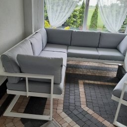 Rafał Macina - Nowoczesny narożnik z białego metalu i szarymi poduchami na tarasie z kostki, widok na ogród przez okno z firankami. Minimalistyczny design i stonowana kolorystyka.