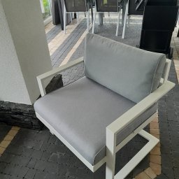 Rafał Macina - Nowoczesny fotel ogrodowy z szarą tapicerką i białą ramą, ustawiony na kostce brukowej. W tle widoczny stół z krzesłami i grill.