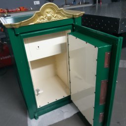 Rafał Macina - Otwarty, zielony sejf z dekoracyjnym, złotym ornamentem na górze, widoczny w warsztacie. Wnętrze sejfu jest jasne, a drzwi uchylone, ukazując mechanizm zamka.