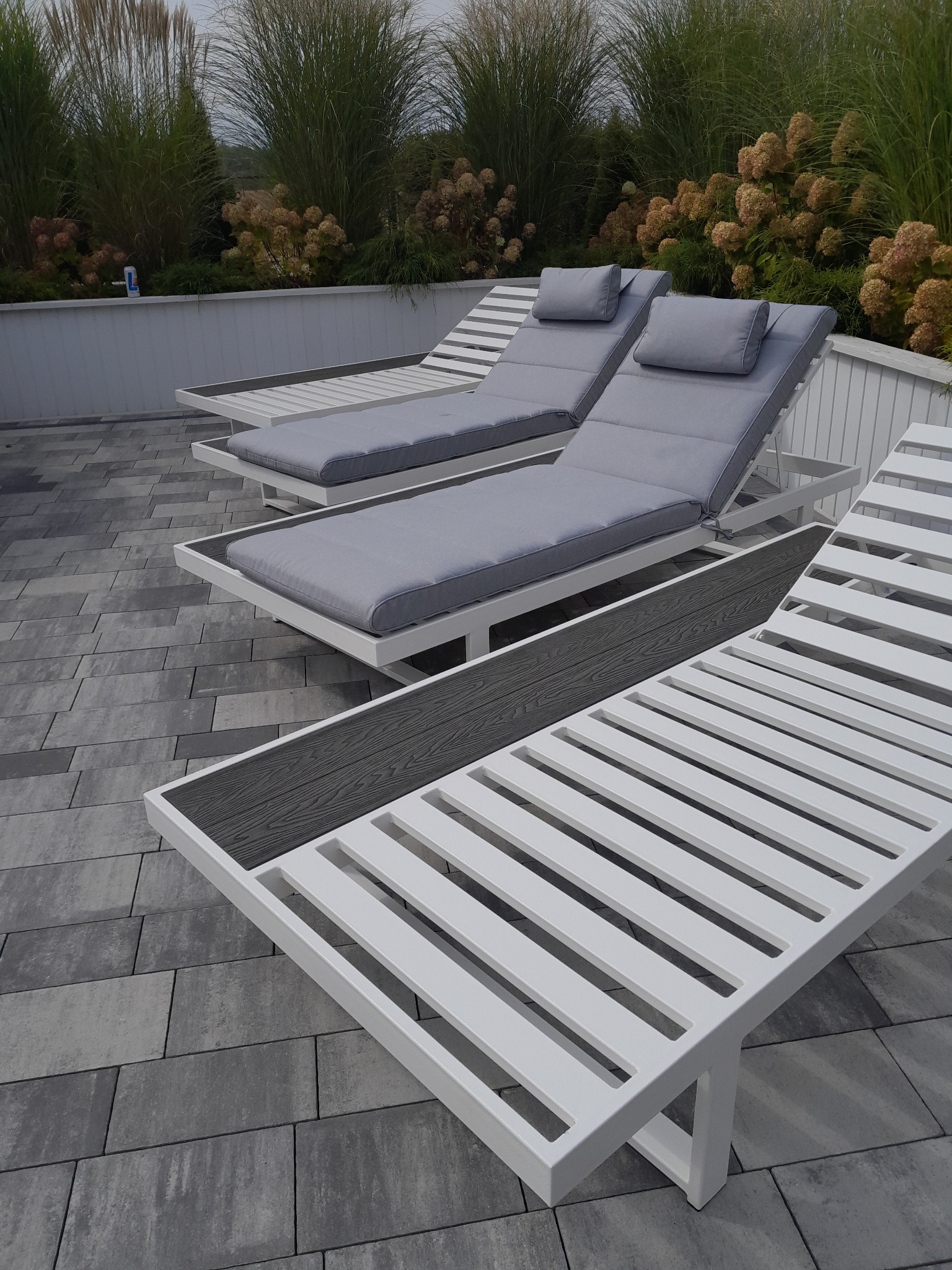 Nowoczesne, białe leżaki z szarymi poduszkami na szarym bruku, otoczone zielenią. Minimalistyczny design i spokojna kolorystyka tworzą relaksującą atmosferę.