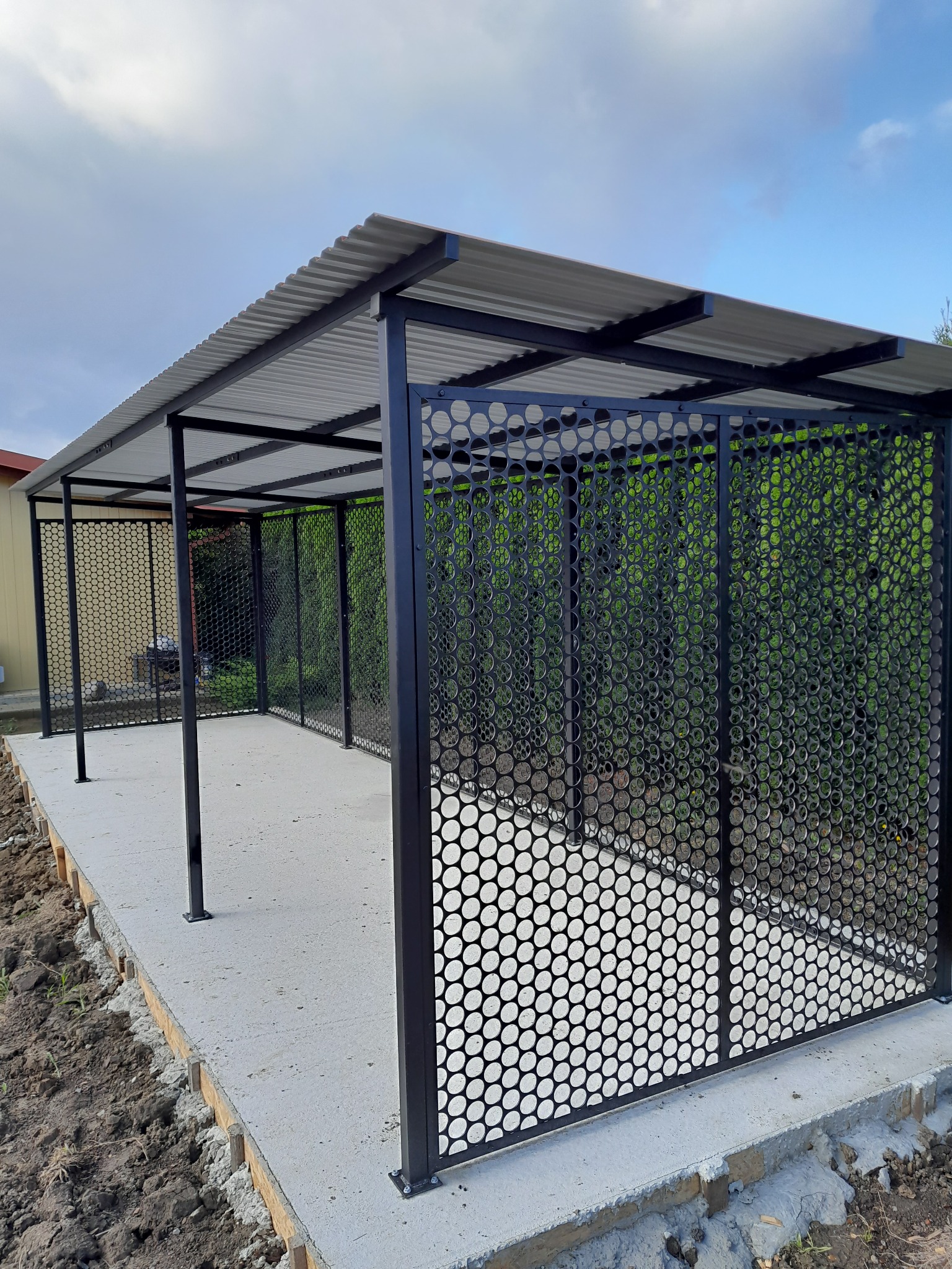 Nowoczesna, czarna konstrukcja z metalu z perforowanymi ścianami i zadaszeniem z blachy falistej, osadzona na betonowej podstawie. Widok z zewnątrz, w tle zieleń.