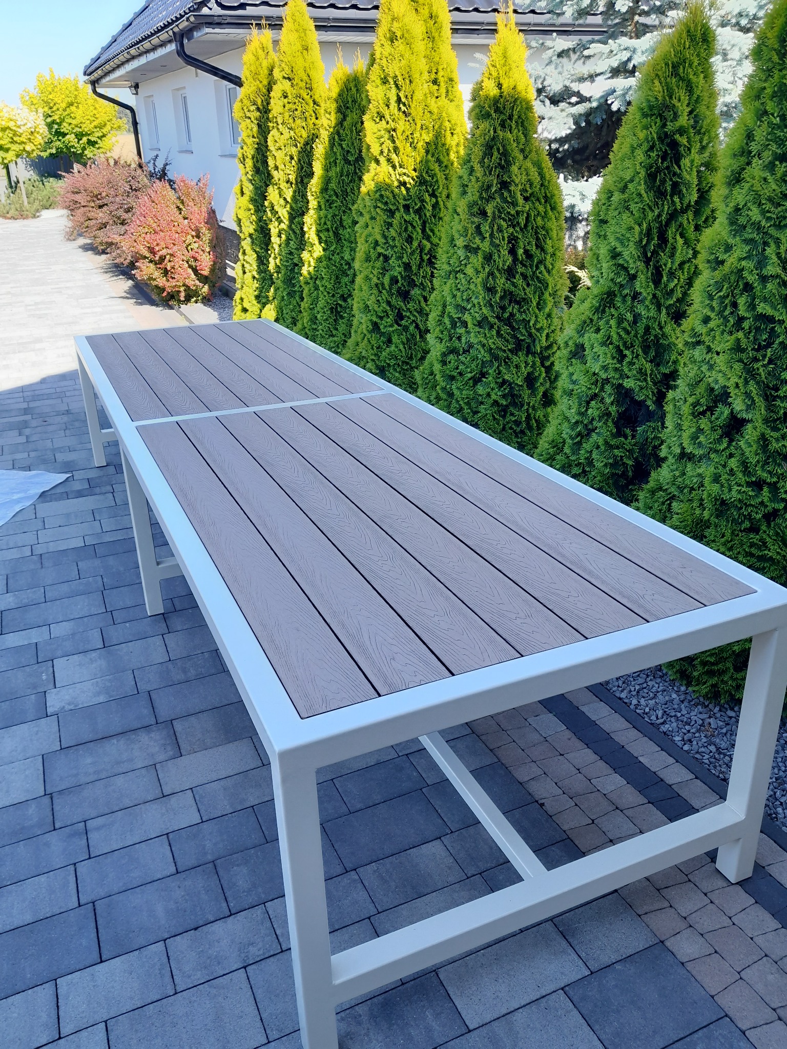 Stół ogrodowy z blatem imitującym drewno, na białym metalowym stelażu, na tle kostki brukowej i zielonych tuj. Nowoczesny design.