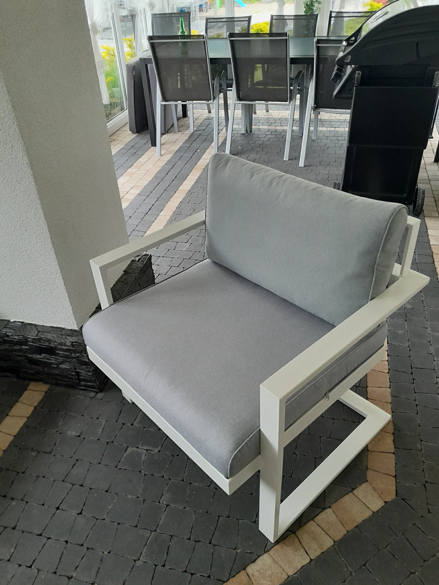 Nowoczesny fotel ogrodowy z szarą tapicerką i białą ramą, ustawiony na kostce brukowej. W tle widoczny stół z krzesłami i grill.