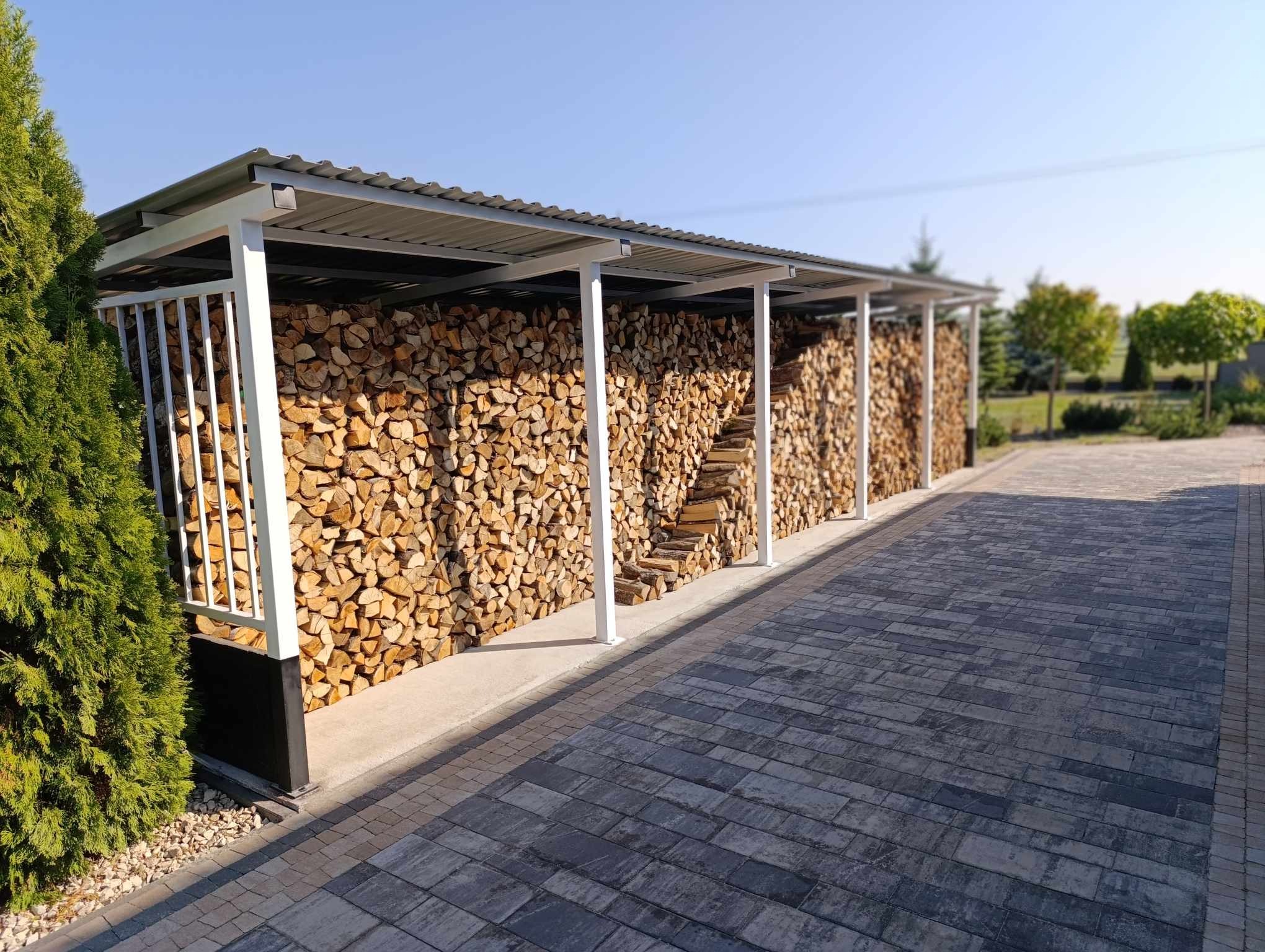 Konstrukcja z metalowym zadaszeniem, wypełniona ułożonym drewnem opałowym. Białe słupy i szara blacha falista. Widok z zewnątrz na kostkę brukową i zieleń.