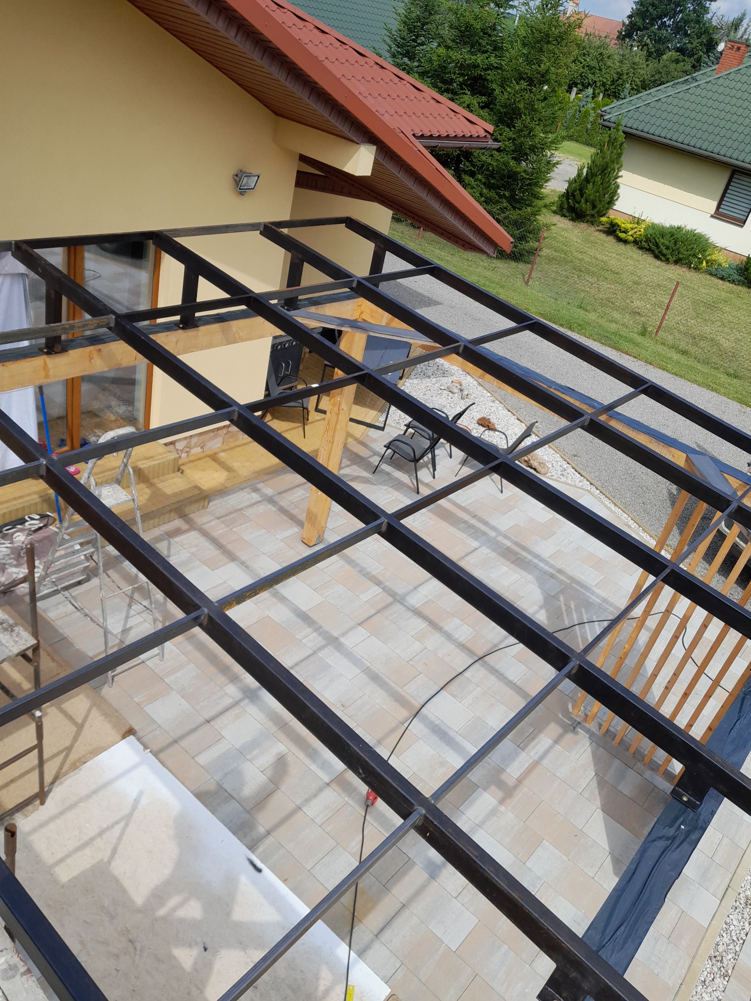 Metalowa konstrukcja z drewnianymi elementami, prawdopodobnie zadaszenie tarasu, widziana z góry. Na pierwszym planie regularna siatka z profili.