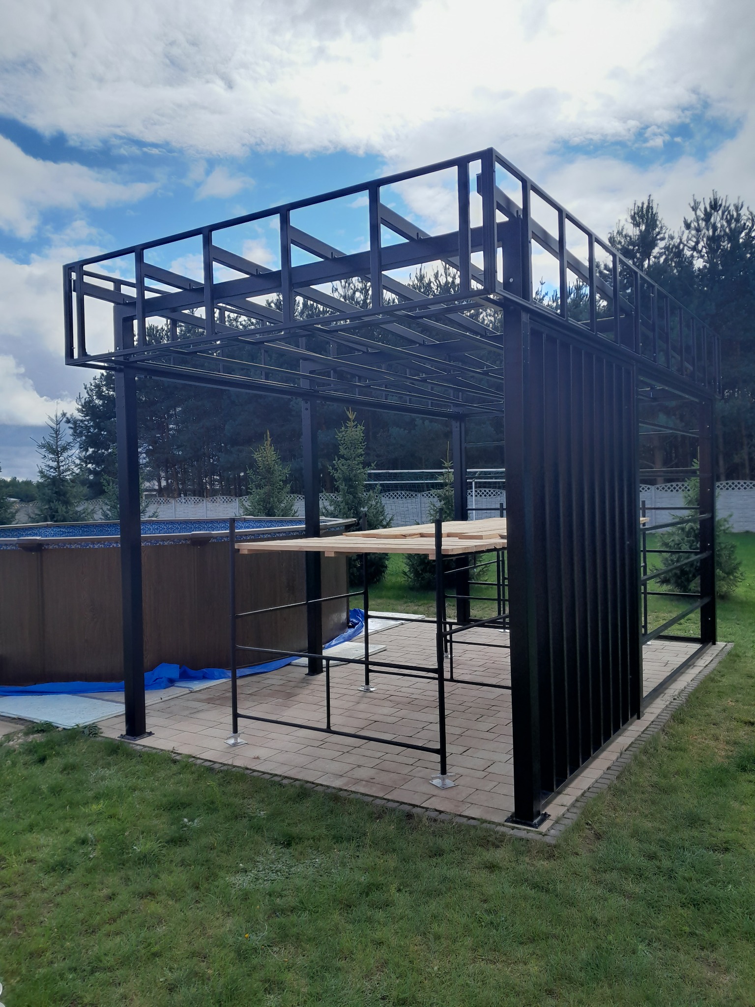 Czarna, metalowa konstrukcja z elementami drewnianymi na zewnątrz, obok basenu. Widoczne niebo z chmurami i trawnik. Konstrukcja w trakcie montażu.