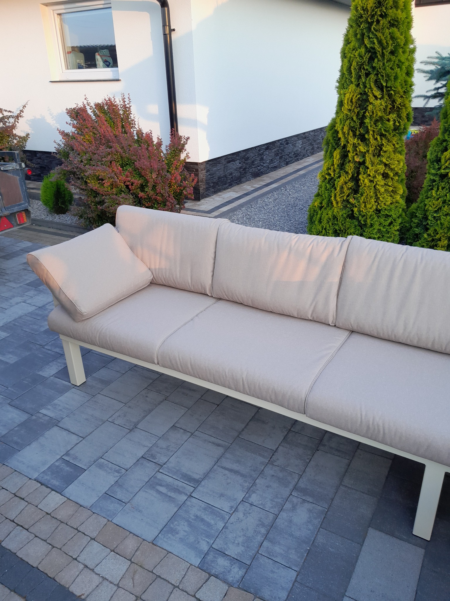 Beżowa sofa ogrodowa z poduszką na kostce brukowej, na tle domu z tujami i krzewami. Sofa z jasnego materiału, ustawiona na zewnątrz.