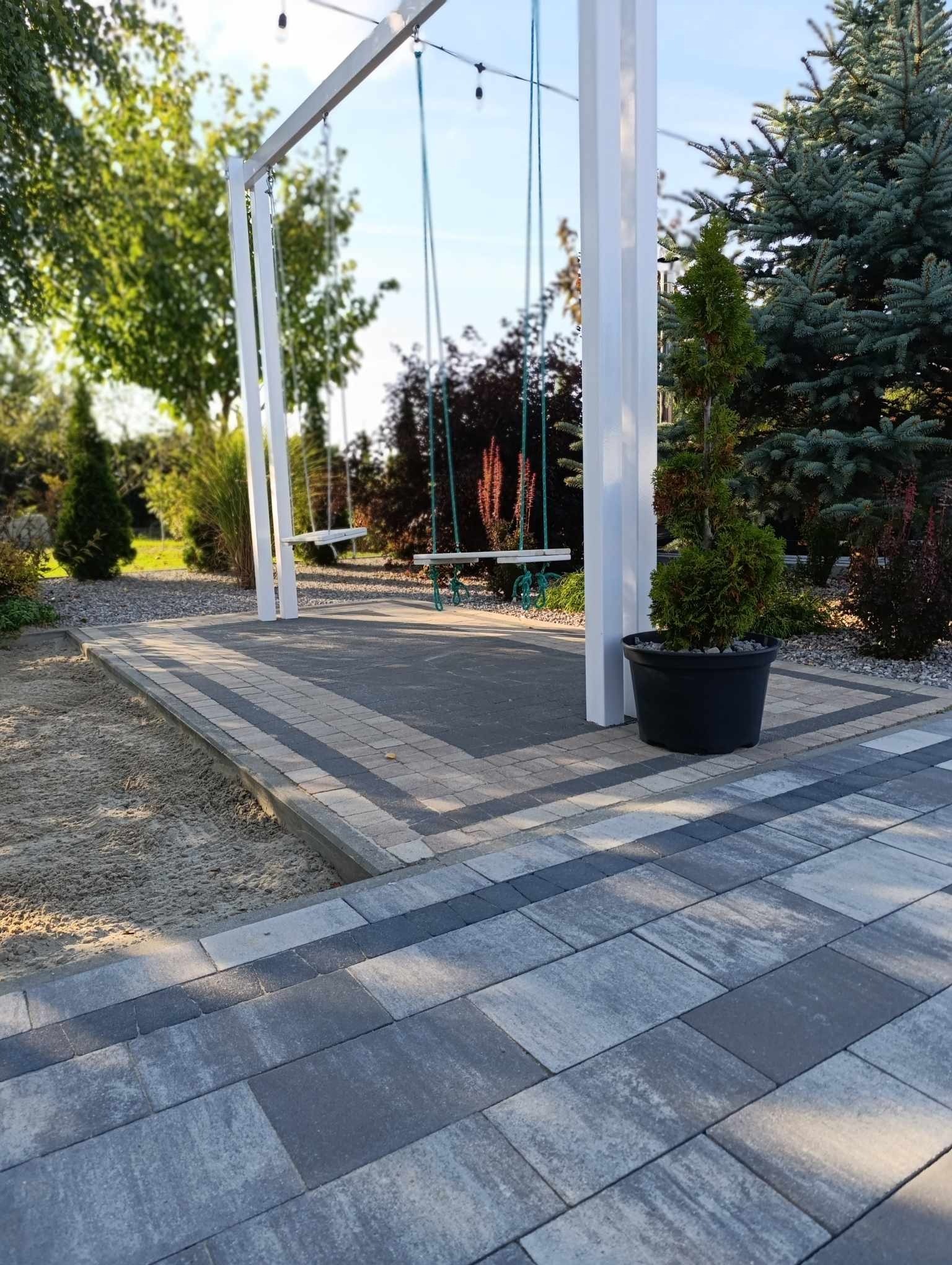 Nowoczesna pergola z huśtawkami w ogrodzie, otoczona kostką brukową i zielenią. Minimalistyczny design, jasne kolory konstrukcji, dekoracyjna roślina w donicy.