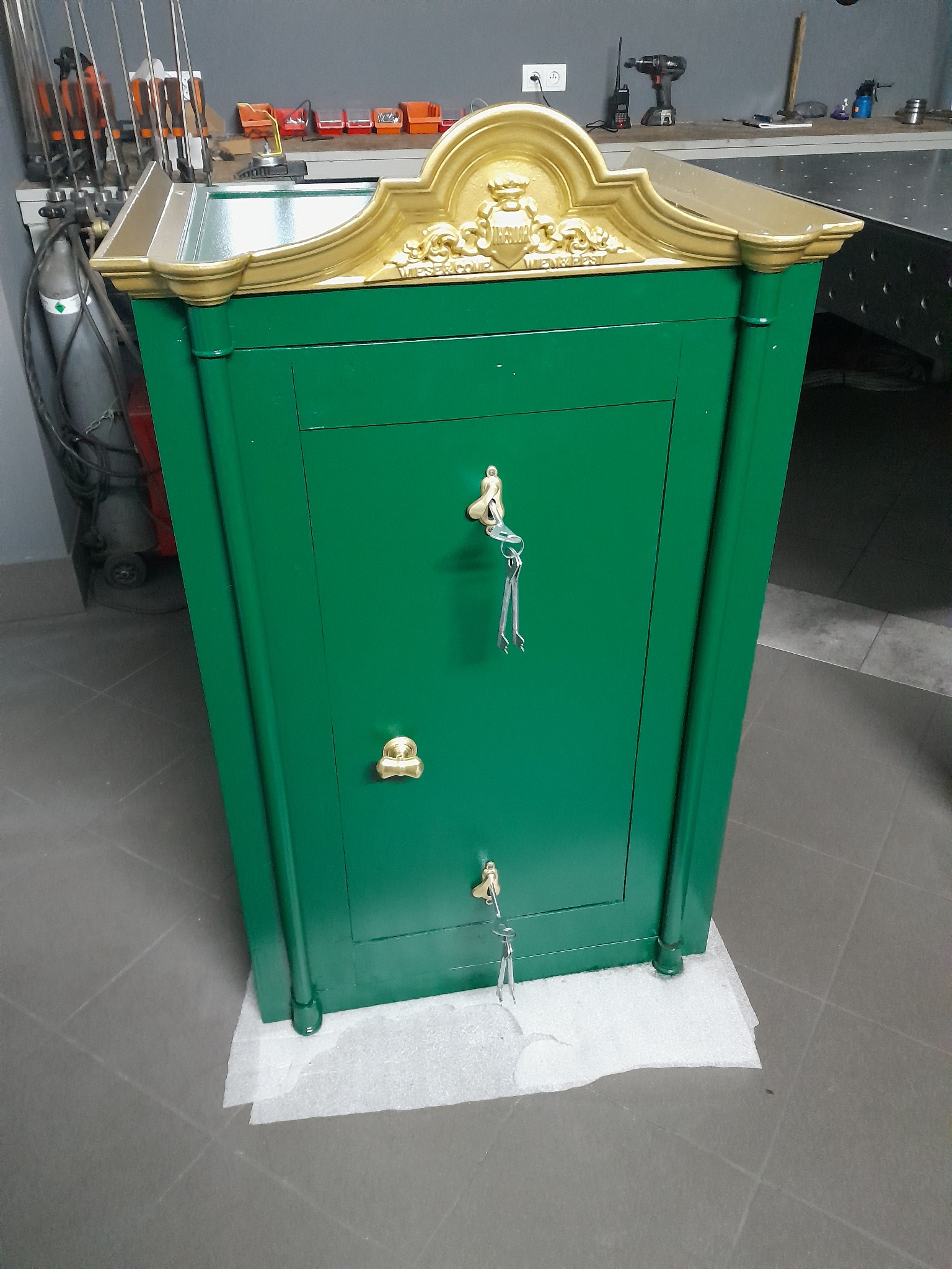 Zielony, metalowy sejf z ozdobnym, złoconym wykończeniem, stojący na podłodze wyłożonej szarymi płytkami. Widoczne narzędzia w tle.