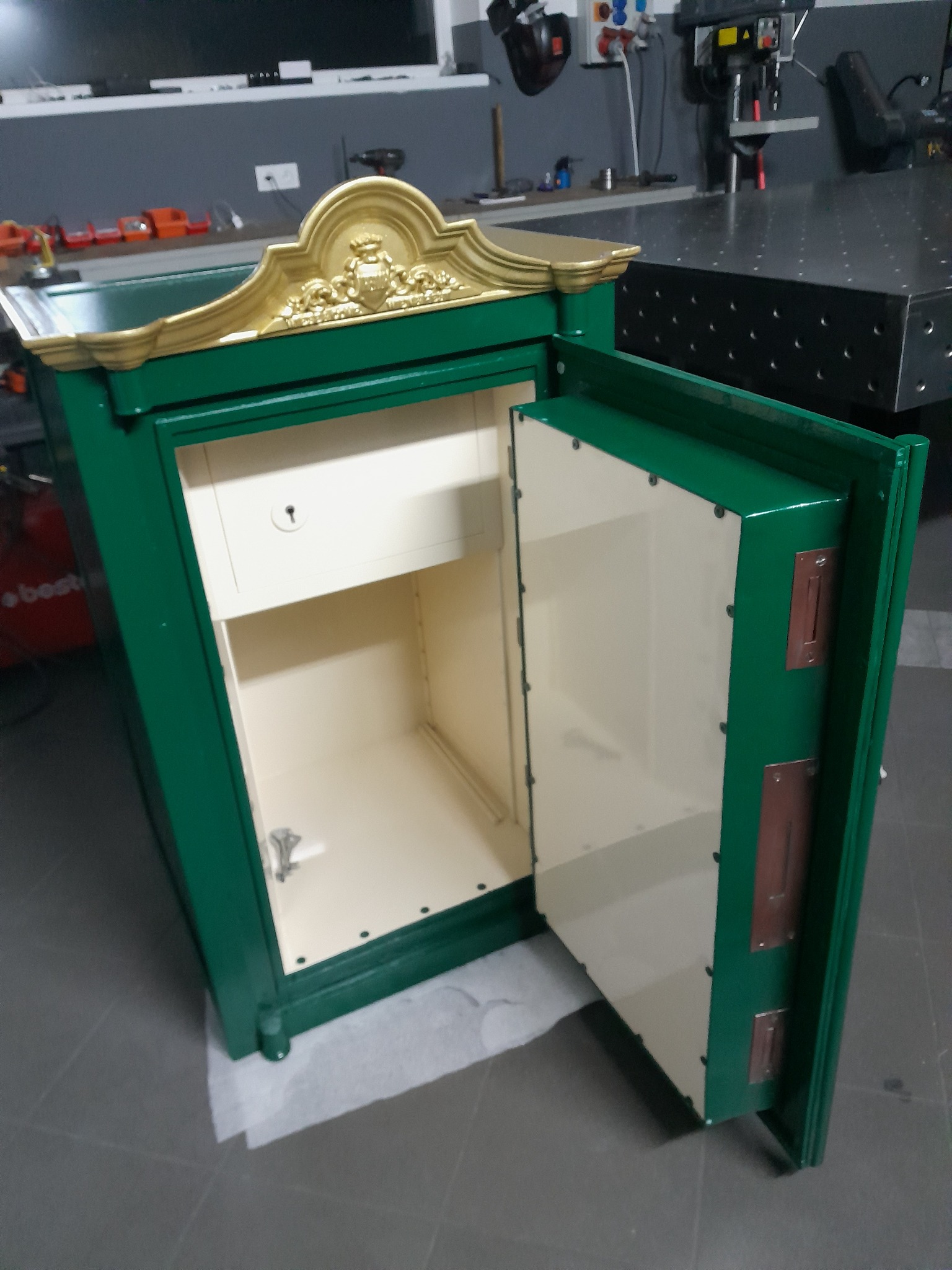 Otwarty, zielony sejf z dekoracyjnym, złotym ornamentem na górze, widoczny w warsztacie. Wnętrze sejfu jest jasne, a drzwi uchylone, ukazując mechanizm zamka.