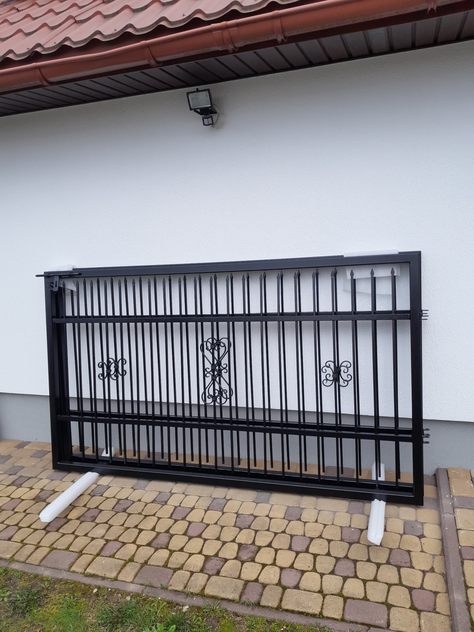 Czarna, metalowa brama z dekoracyjnymi elementami, oparta o białą ścianę i kostkę brukową. Widoczne zabezpieczenia z pianki. Ujęcie z bliska, w dziennym świetle.