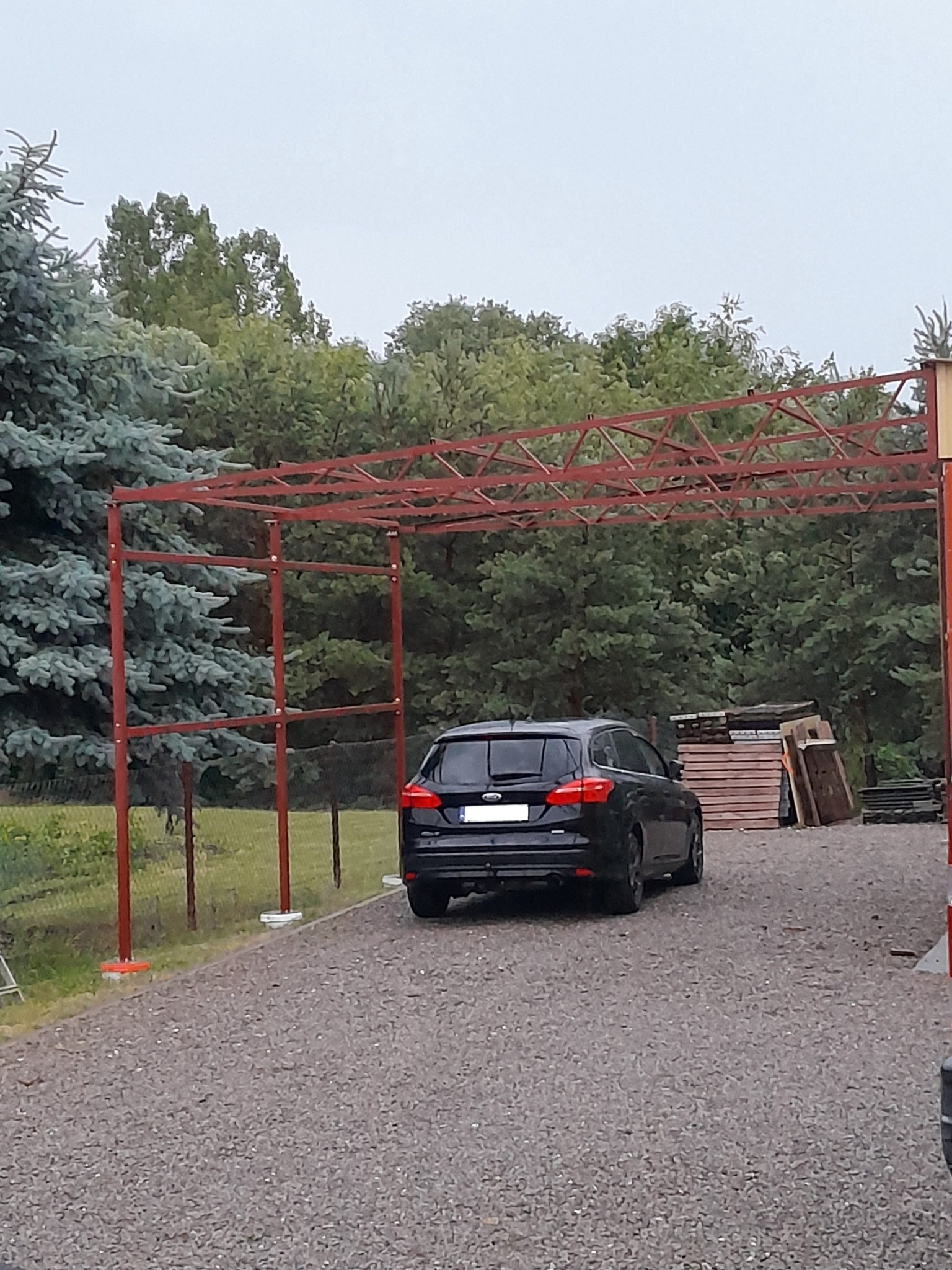 Czerwona, metalowa konstrukcja z kratownicą, prawdopodobnie w trakcie montażu, na tle zieleni i zaparkowanego czarnego samochodu na żwirowym podłożu.