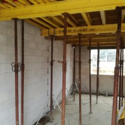 Mi-Mex - Wnętrze budynku w trakcie budowy: ściany z betonu komórkowego, żółte drewniane szalunki stropowe podparte stalowymi stemplami budowlanymi. Widok przez okno na zewnątrz.