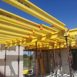 Mi-Mex - Żółte belki stropowe wsparte na stalowych podporach, widoczne białe ściany z bloczków i błękitne niebo. Szalunek stropowy w trakcie montażu na budowie.