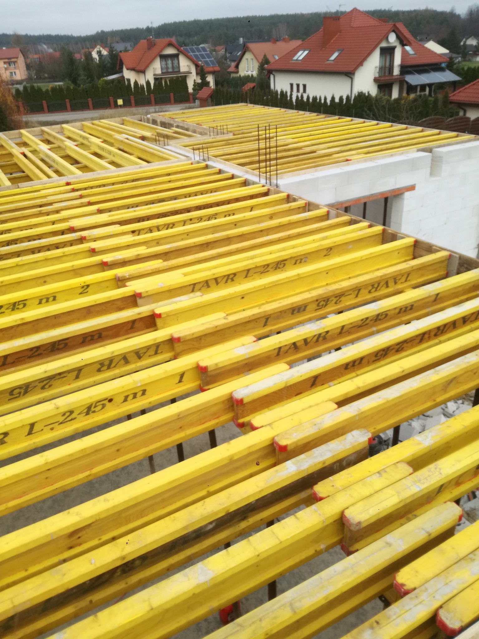 Żółte, drewniane dźwigary stropowe ułożone na budynku w trakcie budowy. Widoczne zbrojenia i bloczki betonowe. W tle zabudowa jednorodzinna i drzewa.