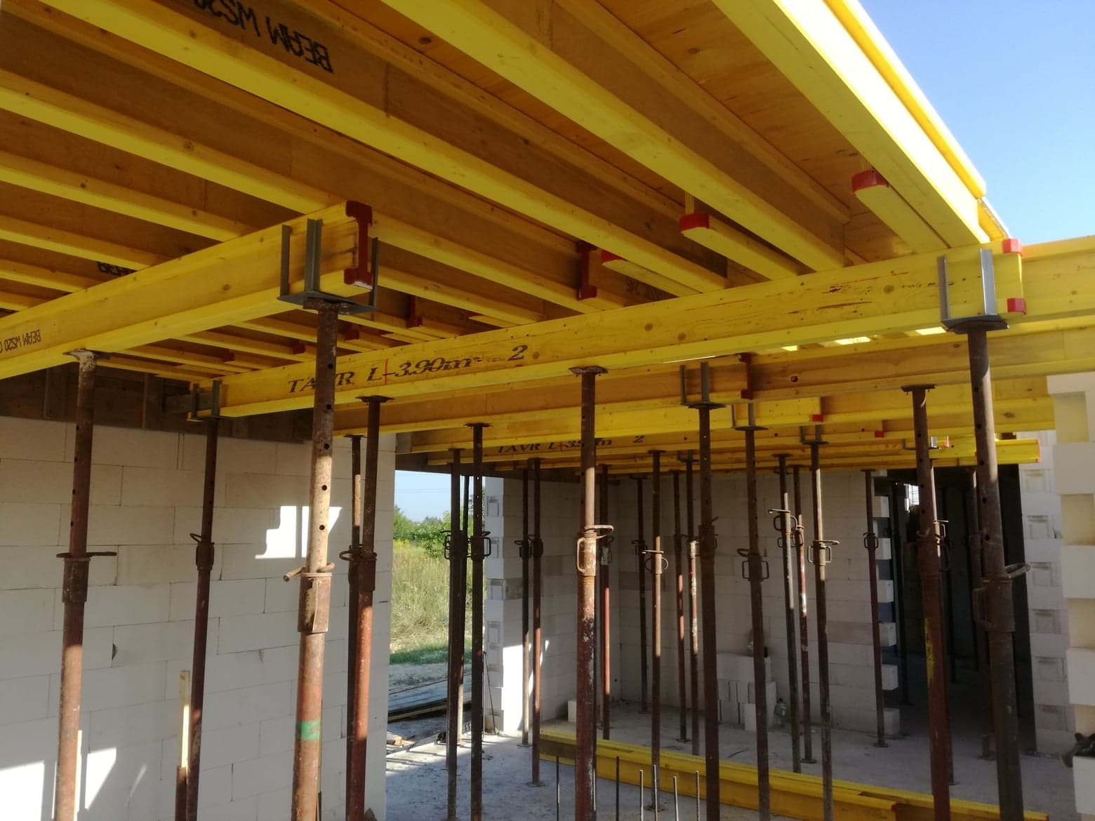 Szczegółowy widok szalunku stropowego z żółtymi belkami i metalowymi podporami w surowym stanie budynku z białych bloczków, ukazujący etap prac fundamentowych.