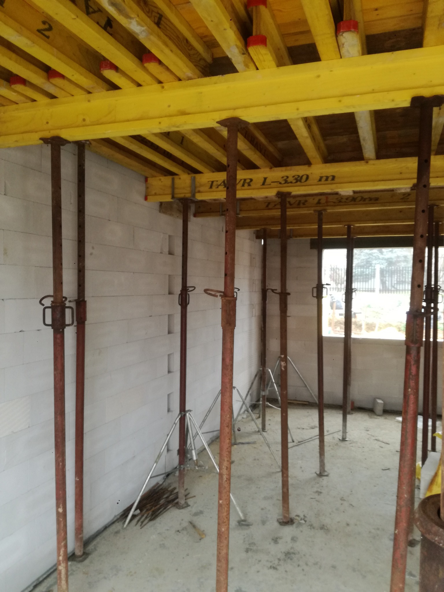 Wnętrze budynku w trakcie budowy: ściany z betonu komórkowego, żółte drewniane szalunki stropowe podparte stalowymi stemplami budowlanymi. Widok przez okno na zewnątrz.