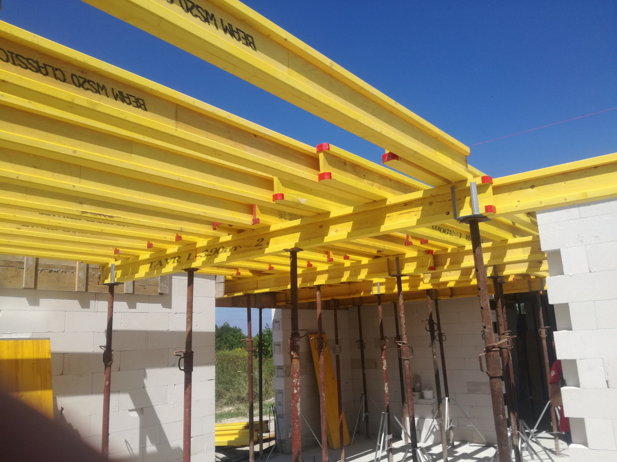 Żółte belki stropowe wsparte na stalowych podporach, widoczne białe ściany z bloczków i błękitne niebo. Szalunek stropowy w trakcie montażu na budowie.
