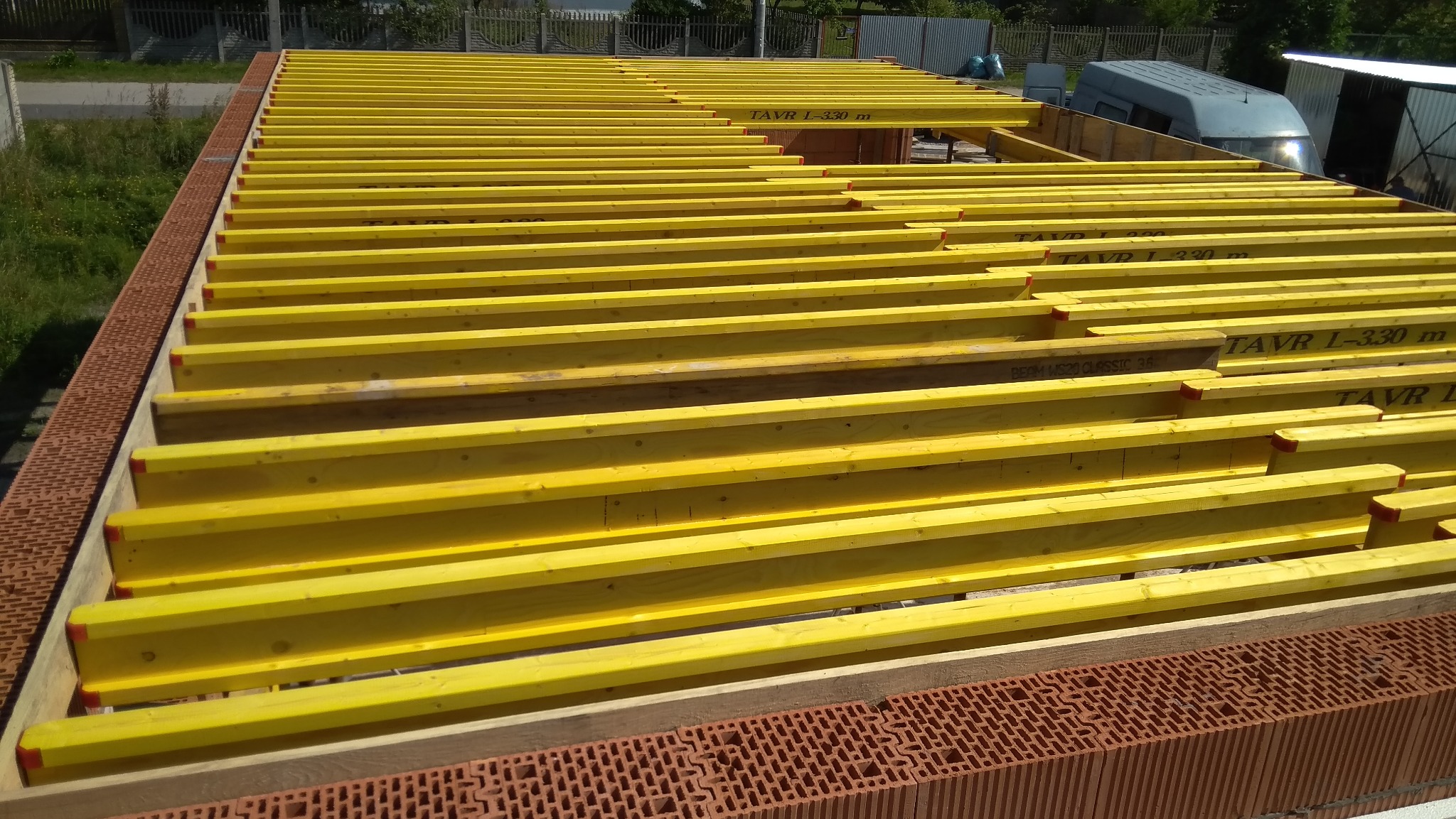 Żółte, drewniane dźwigary stropowe ułożone na murze z czerwonej cegły dziurawki. Widoczne napisy na dźwigarach: TAVR L-330 m, BEAM WS20 CLASSIC 36.