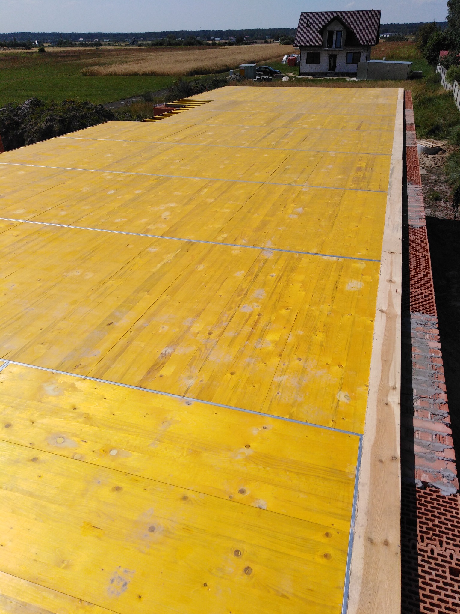 Żółte, drewniane szalunki fundamentów z widoczną konstrukcją z cegieł i deską obrzeżną. W tle dom w budowie i krajobraz.