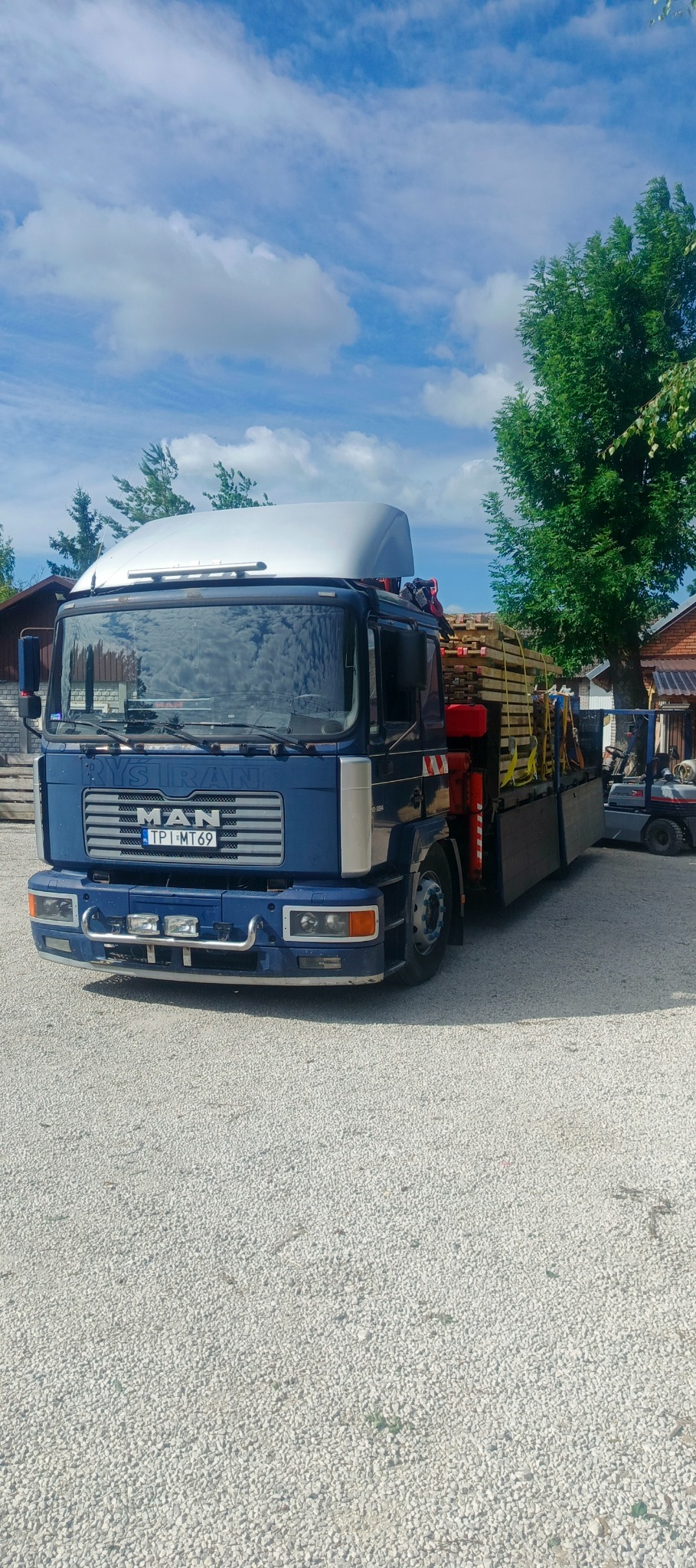 Niebieska ciężarówka MAN z platformą załadowaną drewnianymi elementami konstrukcyjnymi, zaparkowana na żwirowym terenie, w tle budynki i wózek widłowy.