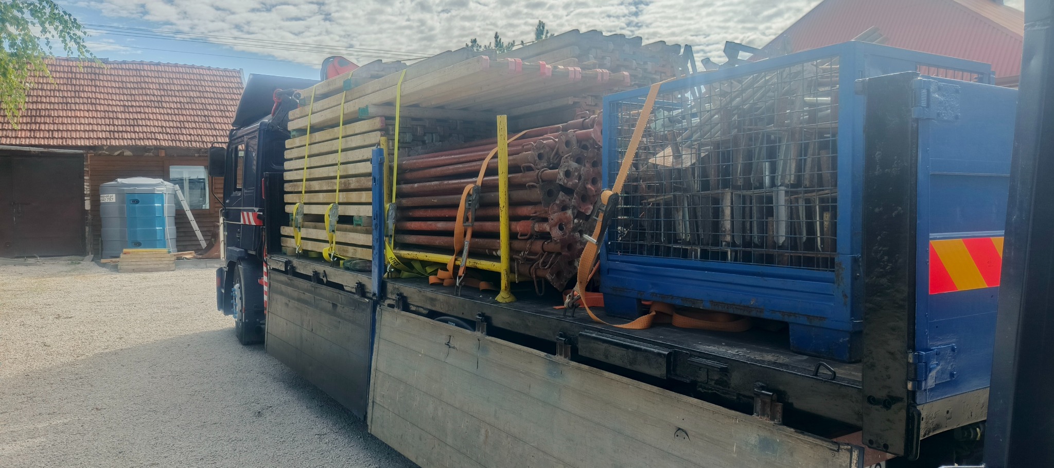 Ciężarówka z platformą załadowana materiałami budowlanymi: drewniane belki, metalowe podpory zabezpieczone pasami transportowymi na tle budynków.