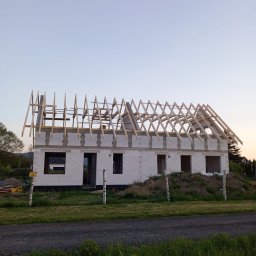 Kamil Grzegolec - Wznoszenie domu z białego bloczka: surowe ściany i drewniana konstrukcja dachu w trakcie budowy, widok z zewnątrz na tle zieleni i błękitnego nieba.