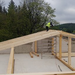 Kamil Grzegolec - Montaż drewnianej więźby dachowej w Jeleniej Górze. Pracownik w kamizelce odblaskowej mocuje elementy konstrukcji na tle lasu i pochmurnego nieba.