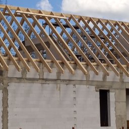 Kamil Grzegolec - Wznoszenie więźby dachowej na budynku z białych bloczków, widoczne niebo i drzewa w tle. Konstrukcja w trakcie budowy, drewniane elementy więźby.