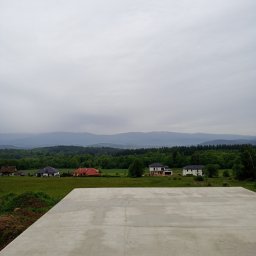 Kamil Grzegolec - Wylana płyta fundamentowa pod budowę domu na tle zielonych wzgórz i lasów. Widok na krajobraz w pochmurny dzień.