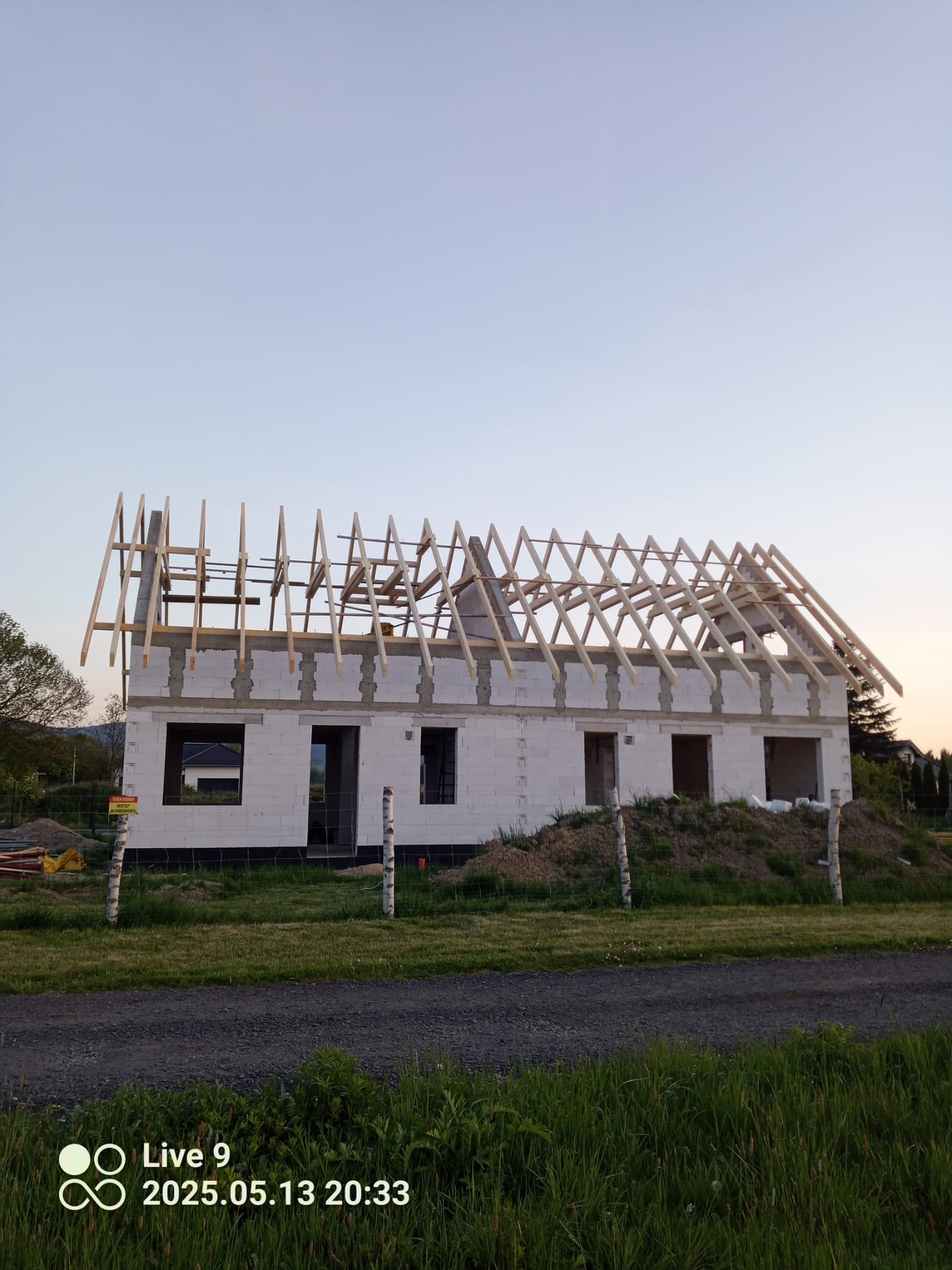 Wznoszenie domu z białego bloczka: surowe ściany i drewniana konstrukcja dachu w trakcie budowy, widok z zewnątrz na tle zieleni i błękitnego nieba.