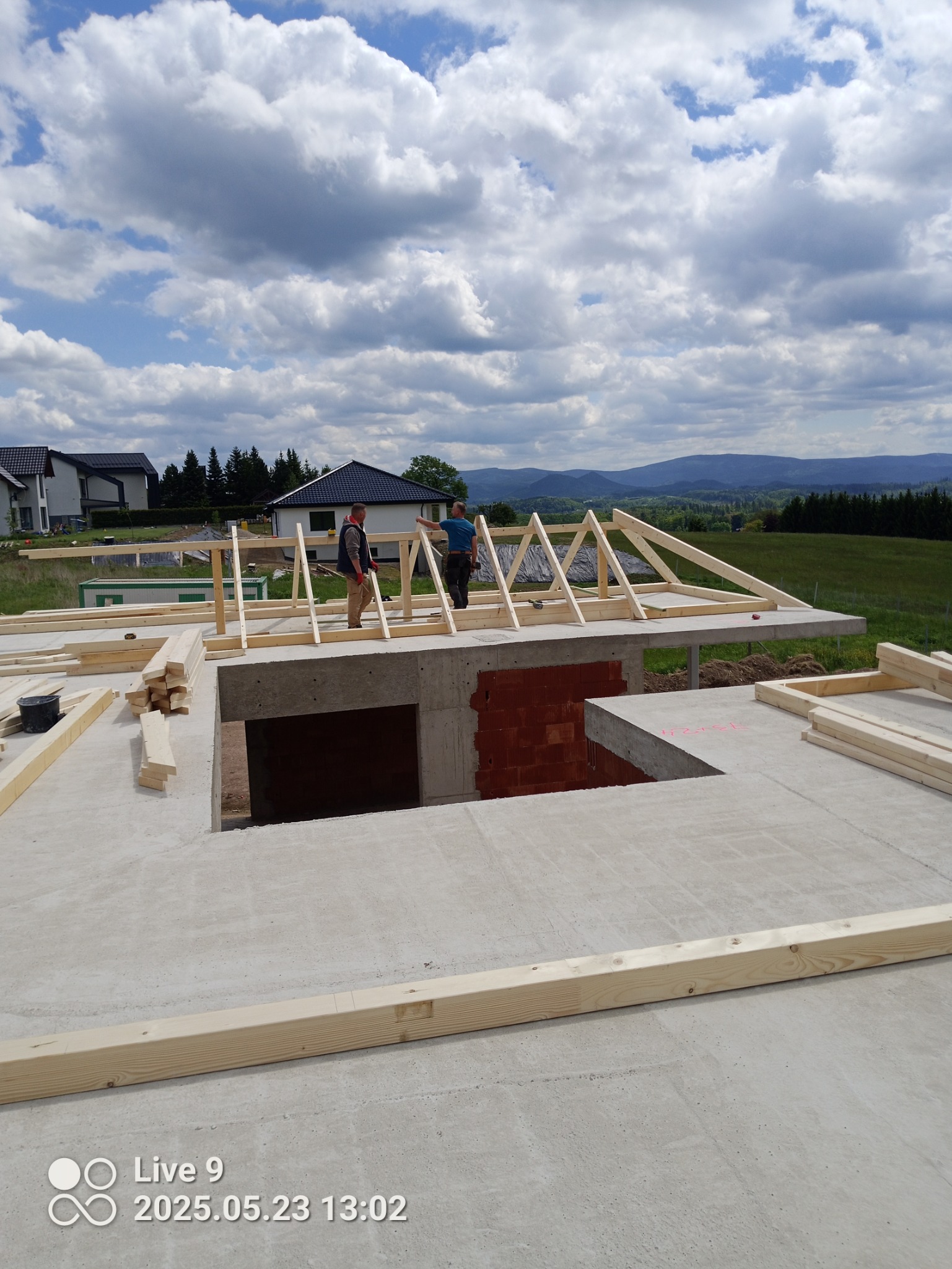 Budowa domu: fundamenty i początki więźby dachowej, dwóch pracowników montuje drewniane elementy konstrukcji na tle malowniczego krajobrazu.