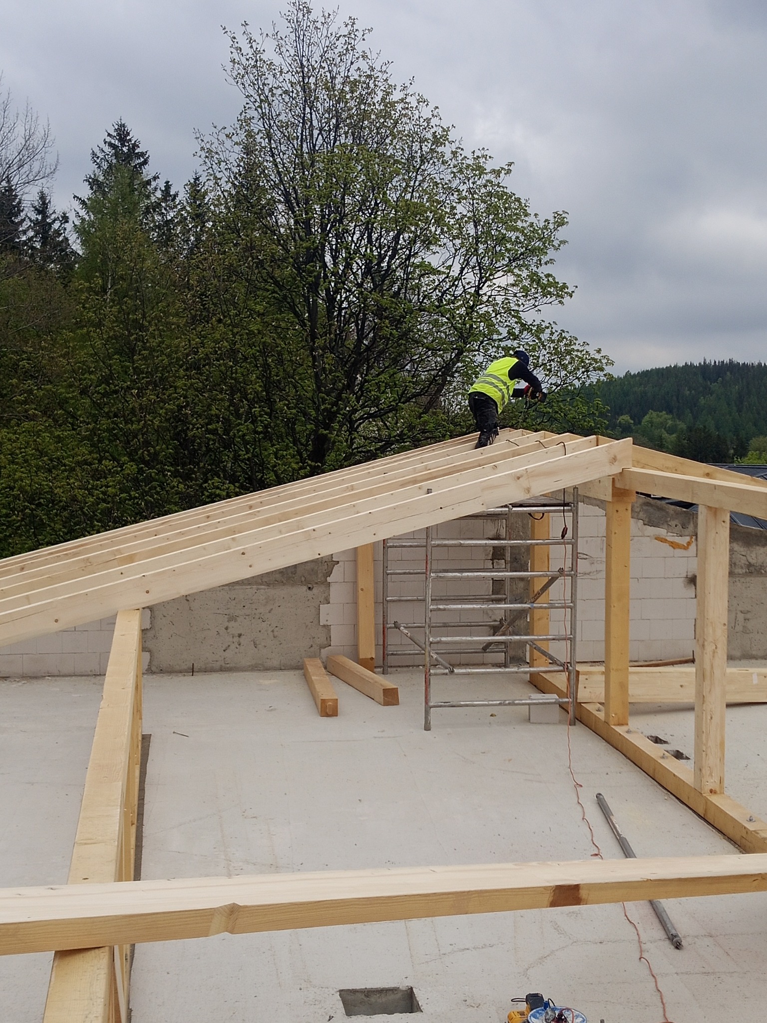 Montaż drewnianej więźby dachowej w Jeleniej Górze. Pracownik w kamizelce odblaskowej mocuje elementy konstrukcji na tle lasu i pochmurnego nieba.